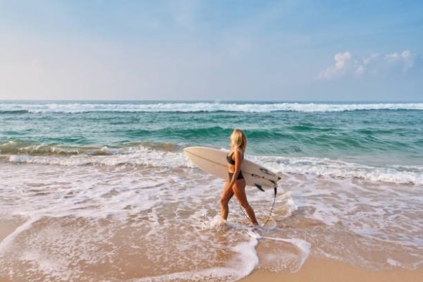 istockphoto-2193451115-612×612 Blonde woman with surfboard walks on sandy beach. Ocean waves crash, sea breeze flows. Active lifestyle and freedom vibe. Surfer girl on sunny day coastal adventure.